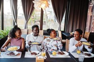 felici amici africani seduti, chiacchierando al bar e mangiando cibo. gruppo di persone di colore che si incontrano al ristorante e cenano. tengono piatti con patatine fritte. foto