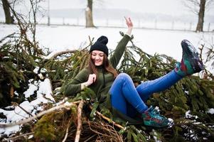 la ragazza indossa una lunga felpa verde, jeans e copricapo nero sui rami del pino nelle giornate invernali. foto