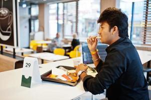 uomo indiano alla moda seduto al fast food contro il suo computer portatile e mangiare patatine fritte. foto