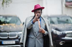 elegante modello uomo afroamericano in cappotto grigio, giacca cravatta e cappello rosso contro due suv nere d'affari e mostra il segno bang bang, tema gangster. foto