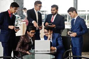 gruppo di sei uomini d'affari indiani in giacca e cravatta seduti in ufficio sul caffè guardando il laptop e bevendo caffè. foto