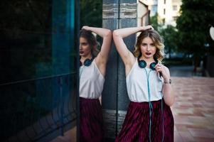 ragazza modello bionda alla moda e bella in elegante gonna di velluto di velluto rosso, camicetta bianca, posata con telefono e auricolari contro le finestre dell'edificio della città. foto