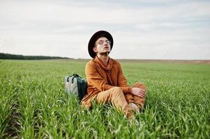 uomo alla moda in occhiali, giacca marrone e cappello con borsa in posa su campo verde. foto