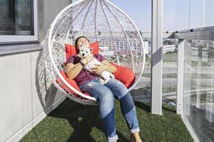 donna millenaria con cucciolo bianco seduto in una moderna poltrona sospesa sul balcone di un condominio e sorriso. foto