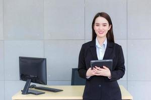 la donna lavoratrice professionista asiatica in un abito nero tiene il tablet nelle sue mani e sorride fiducioso nella stanza dell'ufficio. foto