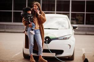 giovane madre con bambino che carica l'auto elettrica presso la stazione di servizio elettrica e parla sul telefono cellulare. foto