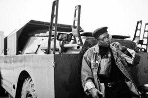 sono il tuo capitano uomo afroamericano in giacca di jeans, berretto e occhiali, fumando sigaro e posato contro un veicolo blindato militare btr. foto