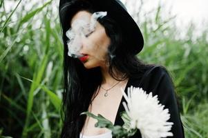 sensuale fumatrice tutta in nero, labbra rosse e cappello. la donna drammatica goth tiene il fiore di crisantemo bianco e fuma sulla canna comune. foto