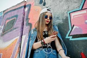 elegante ragazza hipster casual in berretto, occhiali da sole e jeans indossano, ascoltando musica dalle cuffie del telefono cellulare contro il grande muro di graffiti. foto