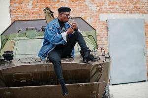 uomo afroamericano in giacca di jeans, berretto e occhiali, accende un sigaro e posa contro un veicolo blindato militare btr. foto