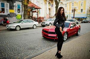 giovane donna riccia e sexy in giacca di pelle contro la muscle car rossa in strada. foto