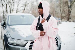 l'uomo afroamericano alla moda in stile urbano in felpa con cappuccio rosa ha posato contro l'auto suv in inverno. ragazzo afro rapper. foto