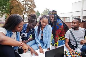 gruppo di cinque studenti universitari africani che trascorrono del tempo insieme nel campus nel cortile dell'università. amici afro neri seduti sull'erba e che studiano con i laptop. foto