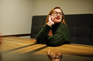 allegra giovane bella donna dai capelli rossi con gli occhiali, indossa un maglione di lana caldo verde, seduto al caffè. foto