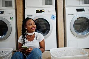 donna afroamericana allegra seduta con gli auricolari e leggere la rivista vicino alla lavatrice nella lavanderia self-service. foto