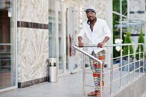 elegante uomo afroamericano in camicia bianca e pantaloni colorati con cappello e occhiali in posa all'aperto. ragazzo nero modello alla moda. foto