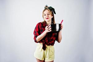 giovane casalinga divertente in camicia a scacchi e pantaloncini gialli pin up style con casseruola e cucchiaio da cucina isolati su sfondo bianco. foto