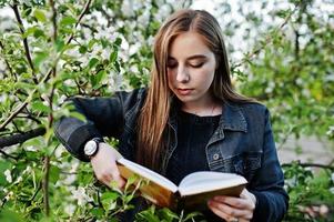 giovane ragazza bruna in jeans contro albero in fiore di primavera leggere il libro. foto