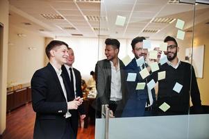 uomo d'affari che punta sul vetro con note di carta colorate. gruppo eterogeneo di dipendenti maschi in abiti formali che utilizzano adesivi. foto