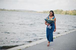 modello afroamericano dalla pelle scura plus size posato in un abito blu lucido con bouquet di fiori contro il lato mare. foto