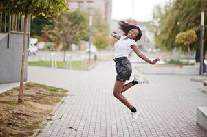 modello slim afroamericano dalla pelle scura posato in pantaloncini di pelle neri e t-shirt bianca. lei salta in aria. foto