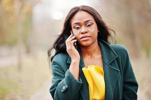 elegante donna afroamericana al cappotto verde e vestito giallo in posa contro il parco autunnale e parlando al telefono. foto