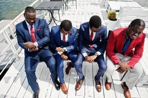 gruppo di quattro uomini di successo felici afroamericani alla tuta. ricchi uomini d'affari neri seduti alle sedie e guardando gli orologi. concetto di tempo lavorativo. foto