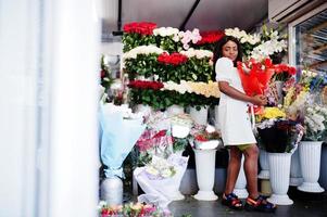 bella ragazza afroamericana in tenero abito bianco con bouquet di fiori in mano in piedi su sfondo floreale nel negozio di fiori fiorista femminile nero. foto