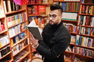 studente arabo alto e intelligente, indossa una giacca di jeans nera e occhiali da vista, in biblioteca con un libro a portata di mano. foto