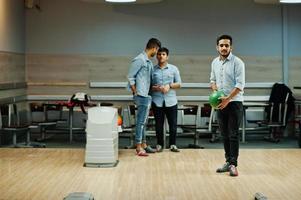 uomo del sud asiatico in camicia di jeans in piedi alla pista da bowling con la palla sulle mani. ragazzo si sta preparando per un tiro. gli amici lo supportano ad alta voce. foto