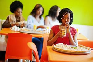 donna africana con pizza seduta al ristorante e bere succo contro ragazze dalla pelle scura. foto