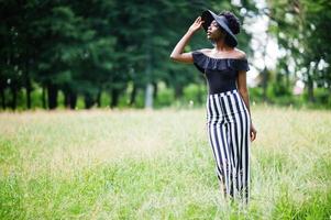 ritratto di splendida donna afroamericana anni '20 in abbigliamento in pantaloni a righe bianche e nere e cappello estivo in posa all'erba verde nel parco. foto