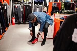 uomo afroamericano casual elegante alla giacca di jeans e berretto nero al negozio di vestiti che prova nuove scarpe da ginnastica rosse. foto