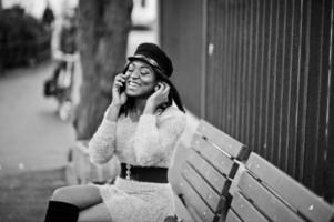 donna afroamericana in abito viola e berretto in posa all'aperto, seduta su una panchina e parlando al telefono. foto