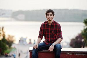 giovane studente indiano in camicia a scacchi rossa e jeans posato in città. foto
