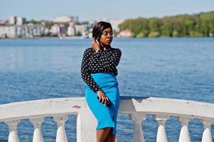 elegante modello afroamericano ragazza con gli occhiali, gonna blu e camicetta nera poste all'aperto sul ponte contro il lago. foto