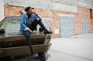 sono il tuo capitano uomo afroamericano in giacca di jeans, berretto e occhiali, fumando sigaro e posato contro un veicolo blindato militare btr. foto