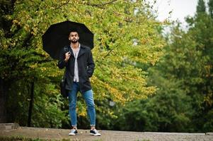 l'uomo alla moda con barba araba alta indossa un cappotto nero con ombrello e custodia per borsa poste in una giornata di pioggia. foto