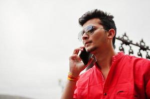 uomo indiano in camicia rossa e occhiali da sole in posa all'aperto e parlando al telefono. foto