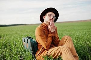 uomo alla moda in occhiali, giacca marrone e cappello con borsa in posa su campo verde. foto