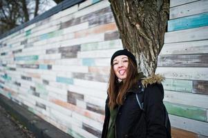 la ragazza indossa un copricapo nero contro la parete di legno mostra la lingua. foto