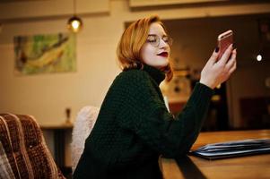 allegra giovane bella donna dai capelli rossi con gli occhiali, maglione di lana caldo verde, usando il suo telefono, mentre era seduto al suo posto di lavoro al bar. foto