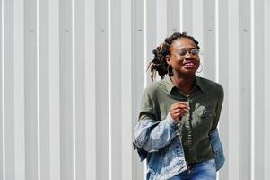 donna africana con i capelli dreadlock, in giacca di jeans e pantaloncini, indossa occhiali posati contro il muro di acciaio bianco. foto