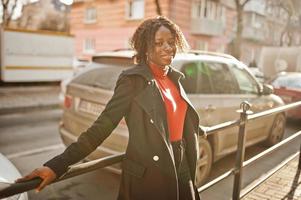 ritratto di una donna africana dai capelli ricci che indossa un cappotto nero alla moda e un dolcevita rosso in posa all'aperto. foto