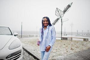 ragazza afroamericana alla moda con dreadlocks che tiene il telefono cellulare a portata di mano, all'aperto contro auto bianca e batterie solari in caso di neve. foto