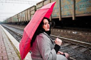 ragazza bruna in cappotto grigio con ombrello rosso nella stazione ferroviaria. foto