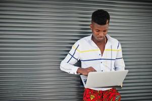 Ritratto di uomo bello ed elegante modello afroamericano in pantaloni rossi e camicia bianca con il computer portatile sulle mani contro lo sfondo dell'otturatore in acciaio. foto