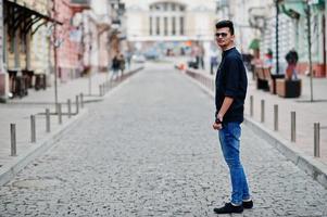 elegante modello indiano uomo in abiti casual, camicia nera e occhiali da sole in posa all'aperto in strada dell'india. foto