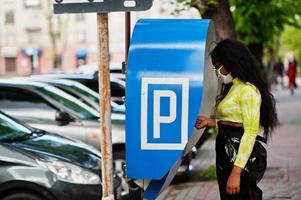 donna afroamericana in posa con maschera facciale per proteggere da infezioni da batteri, virus ed epidemie, utilizzando il terminale della stazione di pagamento del parcheggio. foto