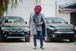 elegante modello uomo afroamericano in cappotto grigio, cravatta giacca e cappello rosso contro due suv nere d'affari. foto